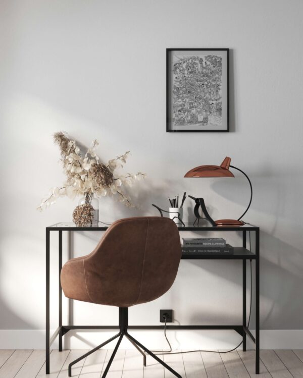 A modern home office workspace featuring a minimalist black metal desk and a cognac-colored leather swivel chair. On the desk sits a curved copper desk lamp with a mushroom-style shade, a vase of dried pampas grass, and a few books. A black-framed botanical art print hangs on the plain white wall above, with soft afternoon sunlight creating shadows across the scene.