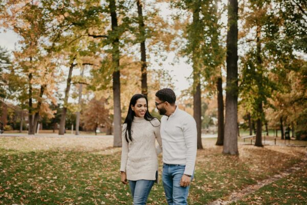 A smiling couple walks together through a park during the autumn season. The woman wears a cream-colored cable-knit turtleneck sweater and jeans, while the man wears a white quarter-zip pullover and blue jeans. They are surrounded by tall trees with orange and yellow leaves, with the ground covered in fallen autumn foliage under soft, diffused natural light.