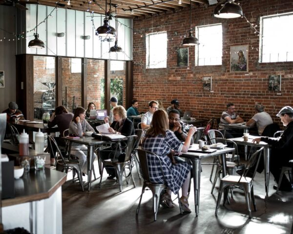 An expansive, industrial-style coffee shop with exposed red brick walls and high ceilings with dark wood beams. The space is filled with patrons seated at metal cafe tables and Tolix-style chairs. Several large, black industrial pendant lights hang from the ceiling, supplemented by festive string lights draped across the room. Large windows allow natural light to stream in, creating high-contrast shadows on the polished concrete floor and highlighting the busy, communal atmosphere.