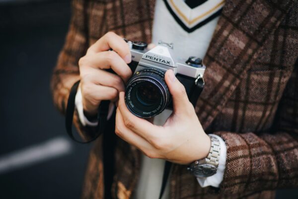 A close-up shot of a person's hands holding a vintage silver Asahi Pentax SLR camera. The individual is wearing a brown plaid wool blazer over a white V-neck sweater with black and gold striped trim. A silver watch is visible on their wrist. The background is a soft-focus dark gray, emphasizing the metallic textures of the camera and the classic, academic style of the apparel.