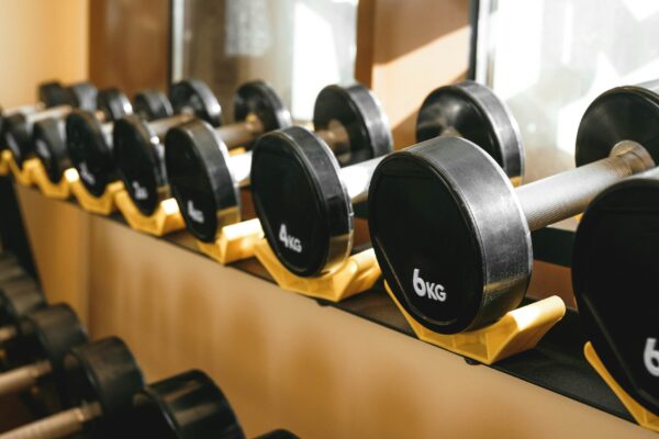 A close-up, angled view of a row of black rubber-coated dumbbells resting on a bright yellow storage rack in a gym. The dumbbells are labeled with their weights, including 4kg and 6kg, and feature textured silver metal handles. The scene is illuminated by bright, clean light reflected in a mirror in the background, emphasizing a high-end and orderly fitness environment.