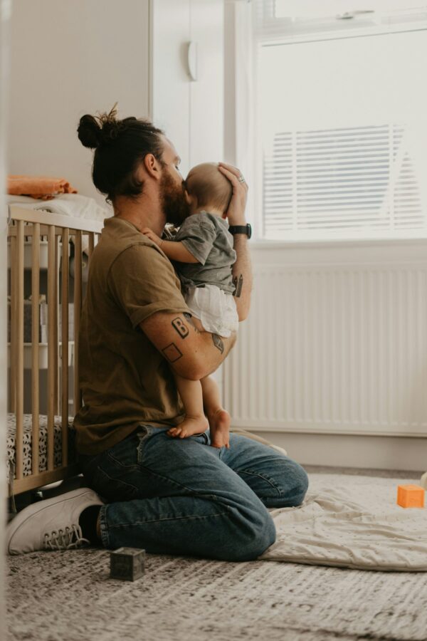 A side view of a father with a beard and hair in a bun, kneeling on a patterned rug in a bright nursery. He is tenderly holding his baby, kissing the infant's forehead. The room is filled with soft natural light from a window with white blinds, highlighting a wooden crib in the background and a white radiator. The father wears an olive green t-shirt and blue jeans, creating a warm, intimate atmosphere of domestic comfort.