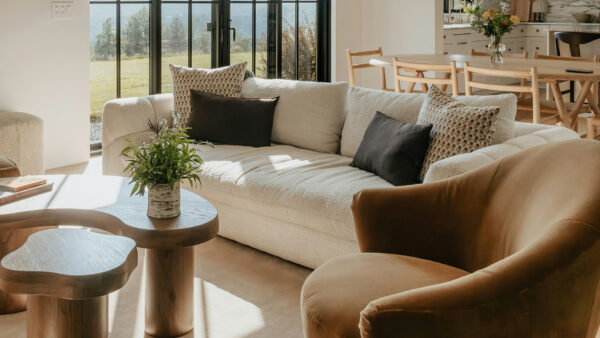 A sun-drenched, modern living room with organic textures. A cream-colored bouclé sofa is styled with dark brown and patterned throw pillows, sitting alongside a velvet cognac-colored armchair. In the foreground, a curved, solid-wood nesting coffee table holds a small potted plant. Large black-framed glass doors in the background reveal a bright, grassy landscape, while a light wood dining set is visible in the adjacent open-concept kitchen space.