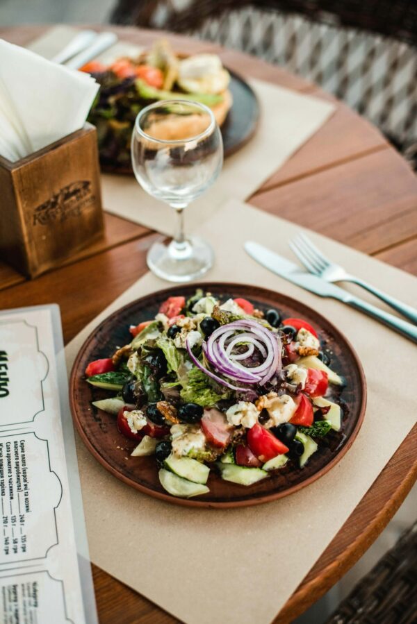 A high-angle shot of a fresh Mediterranean-style salad served on a dark ceramic plate on a wooden cafe table. The salad includes tomatoes, cucumbers, black olives, feta cheese, and is topped with a red onion garnish. In the background, an empty wine glass, a wooden napkin holder, and a menu are visible under soft, warm lighting on a sunlit outdoor patio.