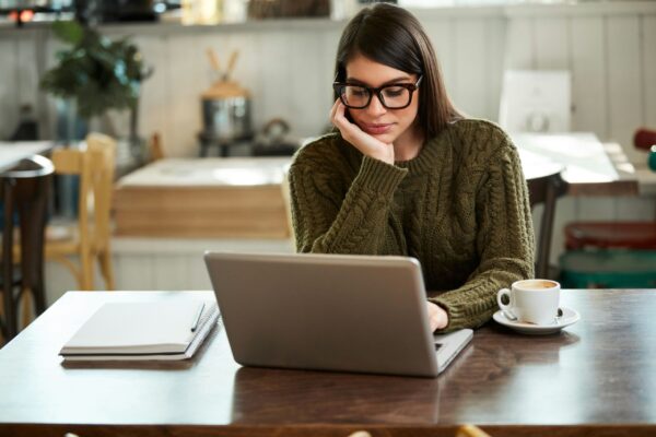 A woman with dark hair and glasses, wearing an olive-green cable-knit sweater, sits at a wooden cafe table. She is focused on a silver laptop with a spiral notebook and a white cup of coffee nearby. The background shows a bright, blurred interior of a modern coffee shop with rustic wooden accents.
