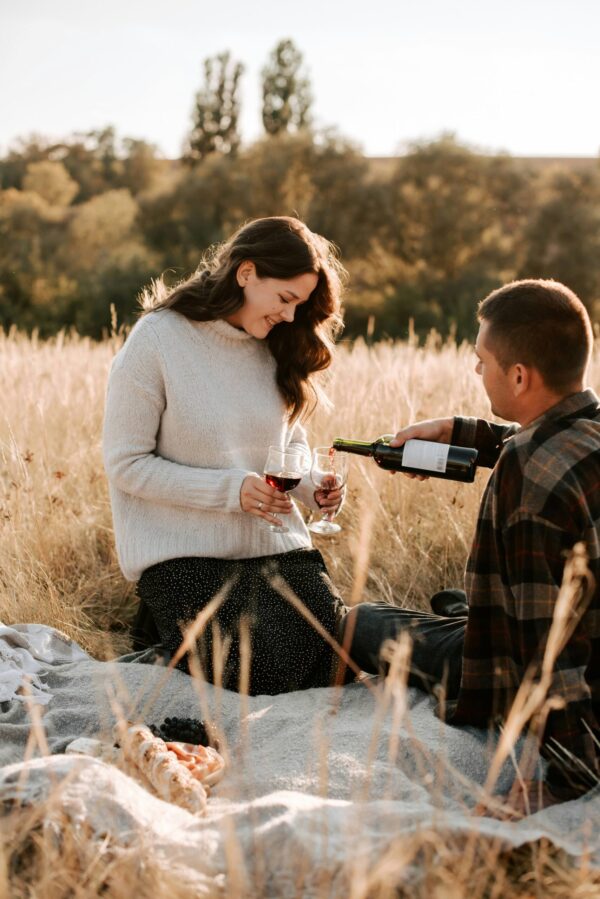 A young couple enjoying a picnic in a field of tall, golden grass during the late afternoon. A man in a plaid shirt is pouring red wine into a glass held by a smiling woman who is wearing a cream-colored sweater and a dark patterned skirt. They are seated on a blanket with a spread of bread and cheese. The scene is bathed in a warm, golden sunset glow, with a soft-focus background of trees and a clear sky.