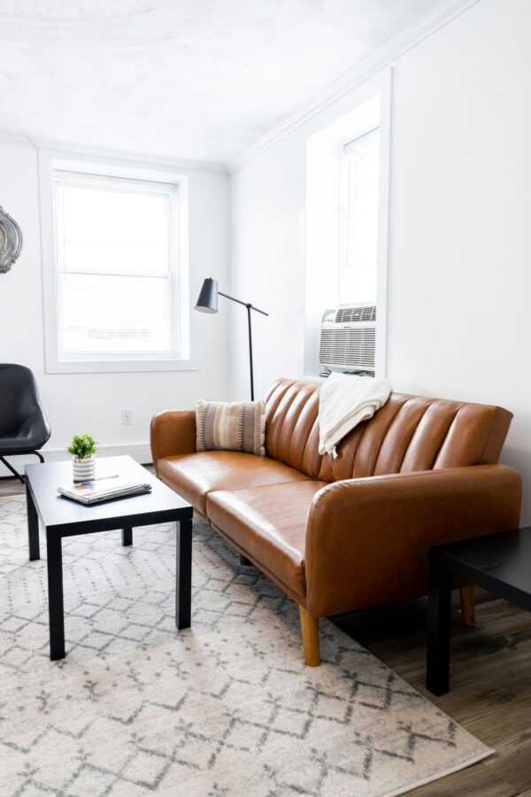 A bright, minimalist living room featuring a cognac leather sofa with vertical stitching and light wood legs. A black articulating floor lamp with a cone-shaped head stands behind the sofa, angled toward a black coffee table. The space is accented by a neutral-toned geometric area rug, white walls, and large windows that flood the room with natural light, creating a clean and airy interior design.