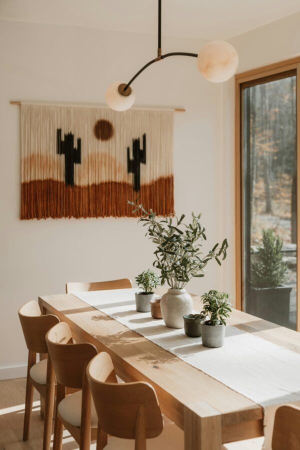 A bright, modern dining room featuring a long oak table with four matching wooden chairs. A contemporary two-arm brass chandelier with frosted globe bulbs hangs above the table, which is decorated with a white linen runner and various potted plants. On the white wall behind the table is a large, desert-themed woven tapestry with a sun and cactus design in rust, cream, and black tones. Large windows to the right let in bright, natural light, highlighting the warm wood grains and organic textures.