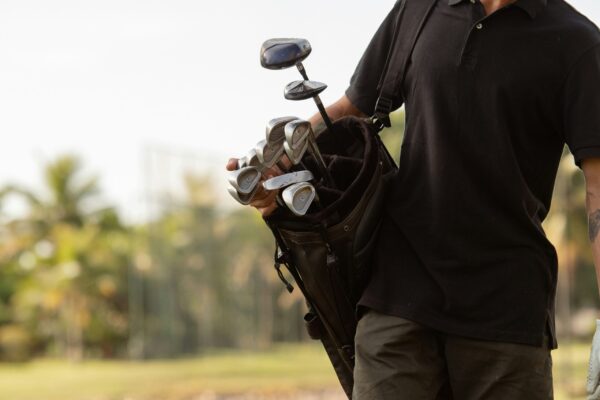 A close-up, mid-section shot of a man wearing a black polo shirt and dark trousers, carrying a premium brown leather golf bag over his shoulder. The bag is filled with various silver-headed golf clubs, including irons and woods. The background features a soft-focus, sunlit tropical landscape with palm trees under a bright sky, highlighting a sophisticated and active lifestyle.