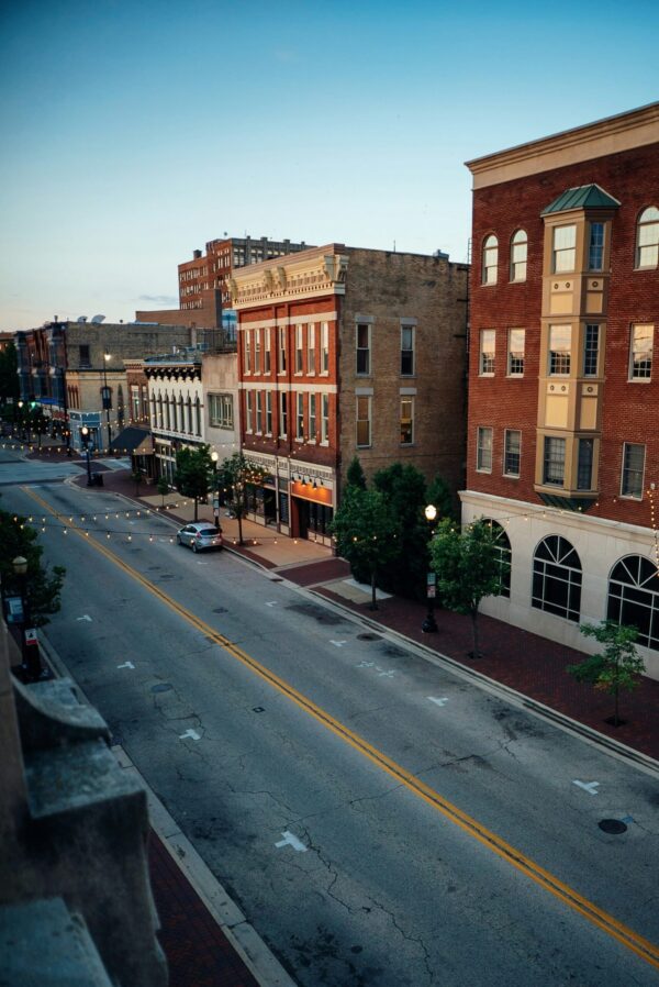 A high-angle, vertical view of a quiet downtown street lined with historic multi-story brick buildings at dusk. Strands of warm white globe string lights are draped across the road between the buildings, illuminating the gray asphalt and yellow lane lines. The architecture features a mix of red and tan brickwork, ornate cornices, and arched windows, set against a clear blue twilight sky.