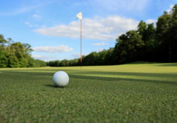 A close-up, low-angle shot of a white dimpled golf ball resting on a vibrant, short-trimmed green putting surface. In the soft-focus background, a tall flagstick with a white flag stands on the green, surrounded by a lush treeline under a bright blue sky with scattered white clouds. The lighting is bright and natural, casting a soft shadow behind the golf ball.